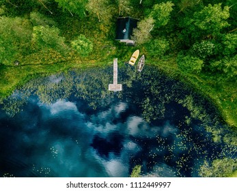 Aerial View Of Forest And Blue Lake With Clouds Reflection In Finland. Sauna House By The Lake Shore. Wooden Pier With Fishing Boats.