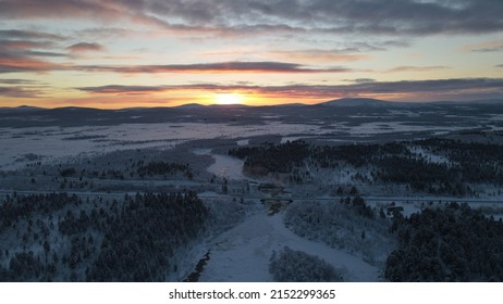 An aerial view of a frozen river flowing through snow-covered forests on a cloudy sunset sky background