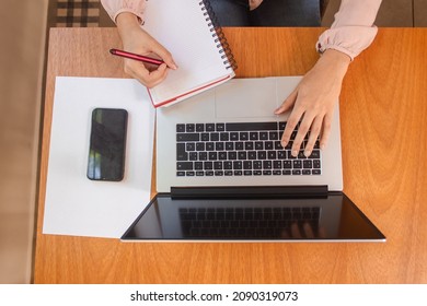 Aerial View Of A Girl's Hands Using A Laptop While Taking Notes In Her Notebook On A Wooden Table.