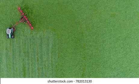 Aerial View Of Green Grass Farm Field With Tractor Making U-turn Raking Grass So It Can Dry Faster And Becomes Dried Grass Or Hay Then Be Picked Up And Kept For Cattle Fodder For Winter Food