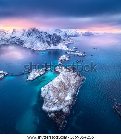 Aerial view of Hamnoy at dramatic sunset in winter in Lofoten islands, Norway. Landscape with blue sea, snowy mountains, rocks and islands, village, buildings, road, bridge, cloudy pink sky. Top view