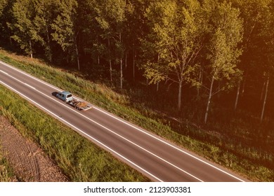 Aerial View Of Hatchback Van With Tilt Car Trailer On The Road Through Green Forest Landscape, Drone Pov Photography