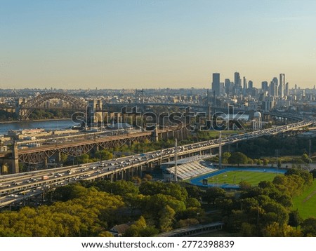 Aerial view of Hell Gate Bridge approaches over Randall's Island sports fields, East River area, and distant Manhattan skyline in clear autumn light.