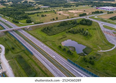 Aerial view of a highway intersection surrounded by green fields and trees. Cars are visible on the road, as well as sound barriers. The image shows a peaceful, rural setting.
