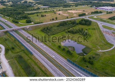 Aerial view of a highway intersection surrounded by green fields and trees. Cars are visible on the road, as well as sound barriers. The image shows a peaceful, rural setting.