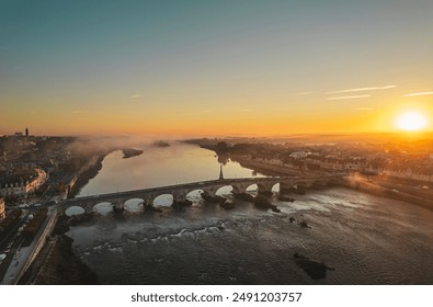 An aerial view of a historic stone bridge in a French city at sunrise, with a golden glow illuminating the scene and a misty river winding through the cityscape - Powered by Shutterstock