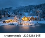 Aerial view of illuminated houses in fairy village in snow, forest, Jasna lake, street lights at winter night. Top view of alpine mountains in fog, snowy pine trees at dusk. Kranjska Gora, Slovenia