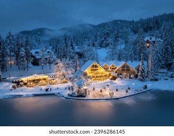 Aerial view of illuminated houses in fairy village in snow, forest, Jasna lake, street lights at winter night. Top view of alpine mountains in fog, snowy pine trees at dusk. Kranjska Gora, Slovenia - Powered by Shutterstock