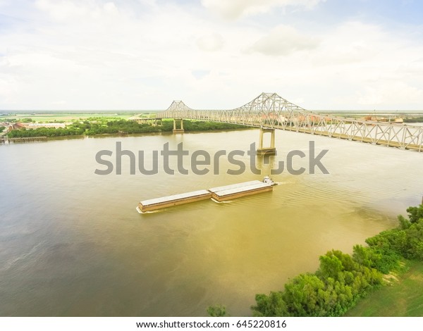 Aerial view of iron cantilever bridge\
over the Mississippi river with tugboat pushing an heavy goods\
barges in rural Louisiana, America. Cloud blue\
sky.