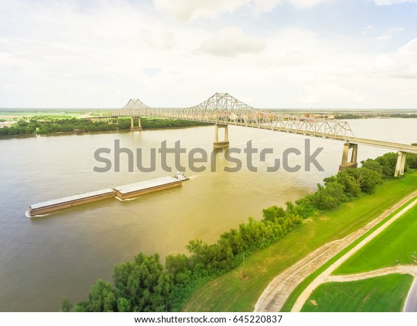 Aerial view of iron cantilever bridge\
over the Mississippi river with tugboat pushing an heavy goods\
barges in rural Louisiana, America. Cloud blue\
sky.