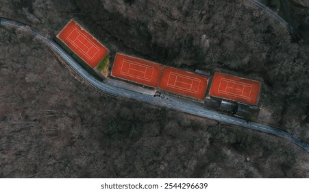 Aerial view of an isolated set of vibrant red tennis courts nestled in a bare, leafless forest, intersected by a winding road, creating a moody, aband - Powered by Shutterstock