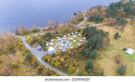 Aerial view of a lakeside campsite with multiple tents arranged in a circle, surrounded by autumn trees and a winding road. The lake is visible on the left, and the campsite is in a forested area. - Powered by Shutterstock