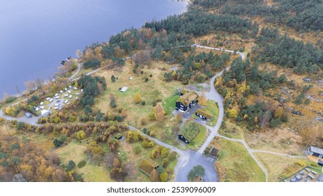 Aerial view of a lakeside campsite surrounded by forested hills. The site includes tents, cabins, and building with green roof. The landscape is dotted with autumn-colored trees, and lake is visible. - Powered by Shutterstock