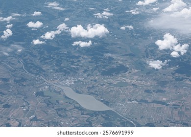 Aerial view of a landscape featuring patchwork fields, a winding river, and a large body of water, with scattered white clouds above and hills in the background