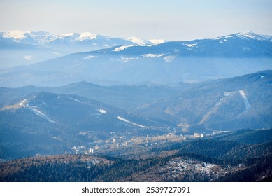 Aerial view of layered mountain ranges partially covered in snow. Valleys filled with dense forests and scattered settlements, while distant snow-capped peaks rise majestically under pale blue sky. - Powered by Shutterstock