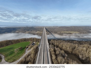 Aerial view of a long highway bridge stretching over a foggy valley with a forest and green fields on either side under a partly cloudy sky - Powered by Shutterstock