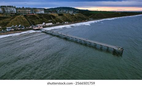 Aerial view of a long pier extending from a coastal town into a dark-blue sea. Buildings line a cliff overlooking the beach and the ocean.   - Powered by Shutterstock