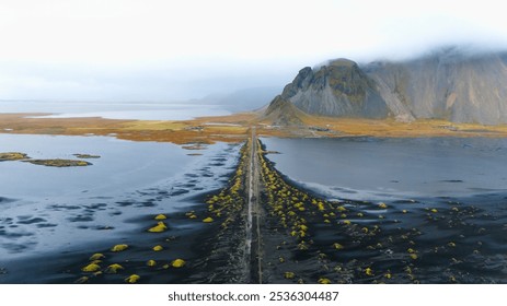 Aerial view of a long road cutting through a surreal coastal landscape in Iceland, with misty mountains in the background and dark sands contrasting with patches of vibrant yellow vegetation. - Powered by Shutterstock