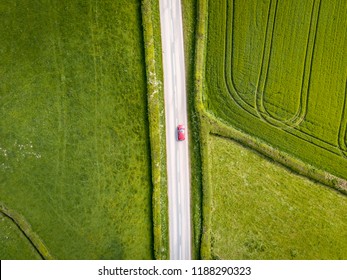 Aerial View Looking Down On A Rural Road In The UK Countryside With A Car Racing Along It. On A Bright Sunny Day, Farmland And Crops Can Be Seen Either Side Of The Road