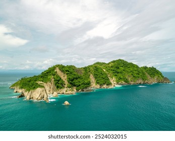 Aerial view of a lush green island surrounded by turquoise ocean waters and rocky cliffs under a cloudy sky. - Powered by Shutterstock