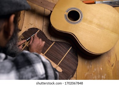 Aerial View Of Luthier's Workshop Table. Craftsman Debating Wood From Back Of Classical Rosewood Guitar With Gouge. Dark Black Background.