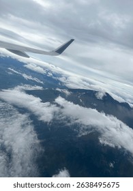 Aerial view of Malang, East Java, seen from an airplane window, showing dramatic clouds, mountain landscape, and aircraft wing under a cloudy sky.