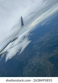 Aerial view of Malang, East Java, seen from an airplane window, showing dramatic clouds, mountain landscape, and aircraft wing under a cloudy sky.