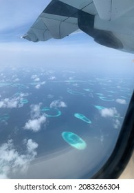 Aerial View Of The Maldives In The Turquoise Water Of The Indian Ocean From The Porthole From Under The Wing Of A Local Airlines Plane Against The Background Of The Sky With Cob Clouds.