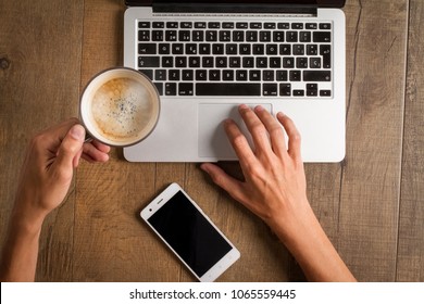 Aerial View Of A Male Hands With A Swatch, Smartphone And A Cup Of Steaming Coffee, Writing On The Keyboard Of A Laptop On A Wooden Table.