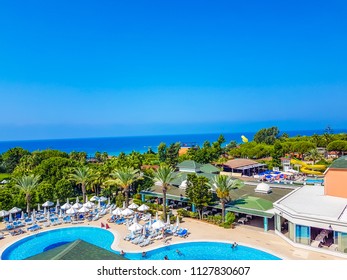 Aerial View Of Mediterranean Sea And Horizon Line And Big Round Swimming Pool With Palm Trees, Restaurant And Sunbeds