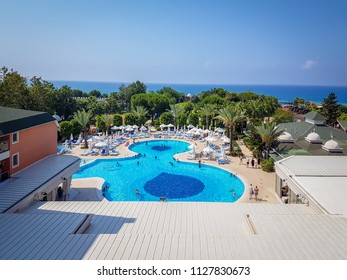 Aerial View Of Mediterranean Sea And Horizon Line And Big Round Swimming Pool With Palm Trees, Restaurant And Sunbeds