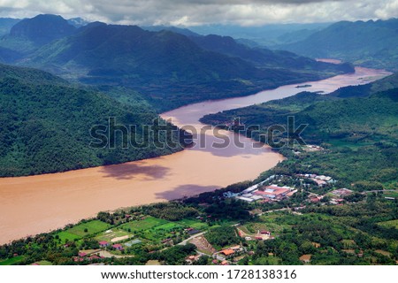 An aerial view of the mighty Mekong River winding near Luang Prabang, showcases the vastness and beauty of the waterway surrounded by lush green mountains and Laos villages.