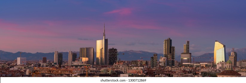 Aerial View Of Milan Skyline At Sunset With Alps Mountains In The Background.