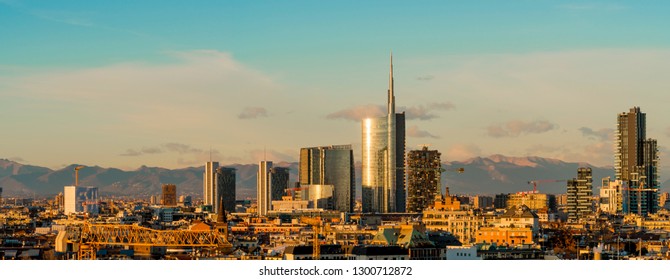 Aerial View Of Milan Skyline At Sunset With Alps Mountains In The Background.