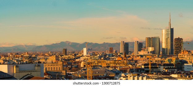 Aerial View Of Milan Skyline At Sunset With Alps Mountains In The Background.