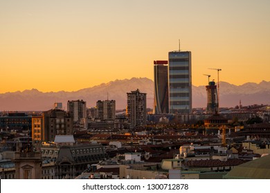 Aerial View Of Milan Skyline At Sunset With Alps Mountains In The Background.