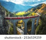 Aerial view of modern red train on Landwasser viaduct in alpine mountains, orange forest at sunset in autumn. Bernina Express, Switzerland. Top view of train in Alps, railroad, colorful trees in fall