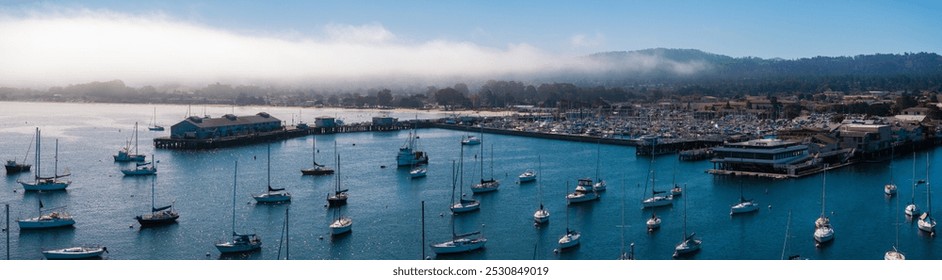 An aerial view of Monterey Harbor, California, shows sailboats in blue waters, a pier extending into the bay, and fog over distant hills. - Powered by Shutterstock