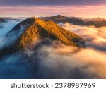 Aerial view of mountain peak in low clouds at sunrise in autumn. Top drone view of hills with red and orange trees in fog, colorful sky in fall. Slovenia. Nature. Mountain valley. Autumn forest. Alps