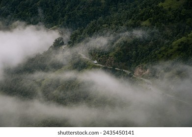 An aerial view of a mountain range enveloped in low-hanging clouds reveals a breathtaking landscape of lush green trees and rocks, with a winding road cutting through it.