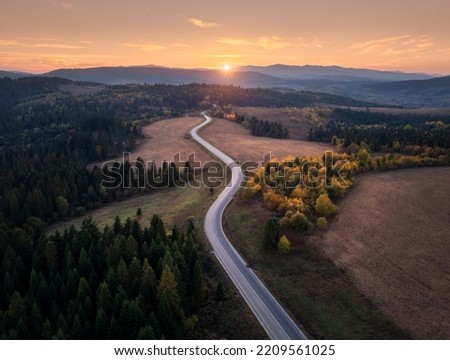 Aerial view of mountain road in autumn forest at sunset in Ukraine. Top view from drone of road in woods. Beautiful landscape with highway in hills, trees, meadows, orange sky, golden sunlight in fall