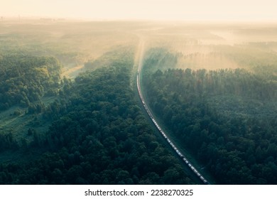 Aerial view of moving freight train in forest. Morning mist landscape with train, railroad, foggy trees. Top aerial drone view near railway station. Train in summer morning forest at fog sunrise. - Powered by Shutterstock