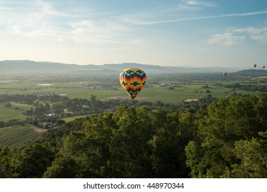 Aerial view of multicolored hot air balloon floating near trees in blue sky above Northern California vineyards