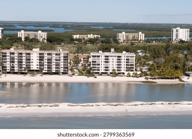 Aerial view of multiple multi-story condominium buildings situated on the white sandy beach of Fort Myers Beach, Florida. The structures feature balconies and large windows overlooking the calm shallow waters of the Gulf of Mexico, with lush green vegetation including palm trees in the foreground and middle ground. This tranquil scene depicts the coastal development prior to the destructive impact of Hurricane Ian in 2022. Fort Myers Beach, USA.