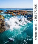 Aerial view of Niagara Falls with turquoise water crashing down surrounded by vibrant autumn foliage. A tour boat approaches the falls, showcasing the grandeur and natural beauty of this iconic landma