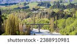 An aerial view of the Old Lower Shotover Bridge in Queenstown, New Zealand, gracefully arching over a tranquil river, framed by mountains and trees as part of the Queenstown trails.