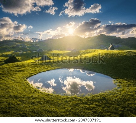 Aerial view of old mountain village on the green hills and pond at sunset in summer. Velika Planina, Slovenia. Top view of wooden houses, meadows, lake, blue sky, clouds. Alpine shepherds? settlement