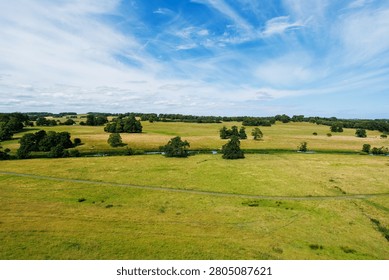 Aerial view of open fields dotted with trees under a bright blue sky, showing an expansive rural landscape featuring hedgerows, small waterways and gentle rolling terrain, United Kingdom