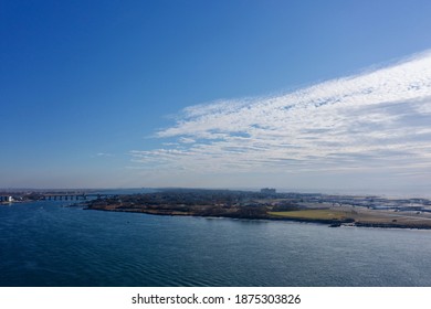 An Aerial View Over East Rockaway Inlet, Looking Towards The Nassau Expressway Bridge. It Is A Sunny Day With Blue Skies And White Clouds. The Waters Are Quiet And Calm.