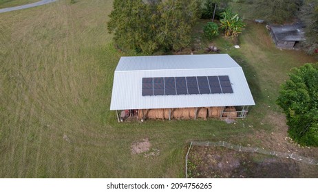 Aerial View Over North Florida Barn Roof With Stacked Hay Bales And Red Tractor With Renewable Energy Solar Panels On Metal Aluminum Roof With Autumn Seasonal Colors Over Farm Garden